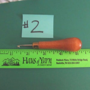 May include: A wooden crochet hook with a metal tip positioned above a green ruler that measures approximately 3.5 inches in length. A light blue paper note with the handwritten text #2 is placed directly above the crochet hook tool.