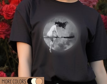 Op de afbeelding: Donkergrijs t-shirt met een zwart-wit afbeelding van een naakt persoon die op een bezem rijdt voor een volle maan. Het shirt heeft de tekst "MORE COLORS" en "100% ORGANIC COTTON" in goud.