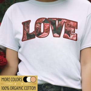 Könnte beinhalten: Weißes T-Shirt mit dem Wort "LOVE" in einem bordeauxroten Umriss, gefüllt mit einem floralen Rosenmuster. Das Shirt hat auch eine kleine Grafik am unteren Rand mit dem Text "MORE COLORS" und "100% ORGANIC COTTON".
