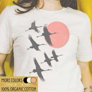 Op de afbeelding: Wit T-shirt met een ontwerp van vliegende kraanvogels in silhouet tegen een roze zon. Het shirt bevat de tekst "MORE COLORS" en "100% ORGANIC COTTON" in een gouden rechthoek. De kraanvogels zijn donkergrijs.