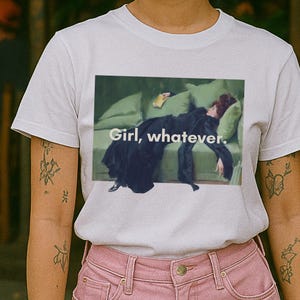 Könnte beinhalten: Ein hellgraues Kurzarm-T-Shirt mit einem rechteckigen Grafikdruck einer Person in einem schwarzen Kleid, die auf einem grünen Sofa liegt und ein Buch hält, mit dem weißen Text "Girl, whatever." in der Mitte des Bildes.