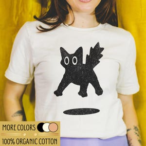 Könnte beinhalten: Cremefarbenes T-Shirt mit einem schwarzen Katzen-Grafik. Die Katze hat große weiße Augen und einen stacheligen Schwanz. Der Text "MORE COLORS" und "100% ORGANIC COTTON" ist unten auf dem Shirt aufgedruckt.
