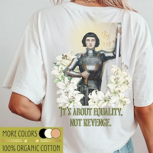 Könnte beinhalten: Weißes T-Shirt mit einer Grafik einer Person in Rüstung, umgeben von weißen Lilien. Der Text "It's about equality, not revenge" steht darunter. Das Shirt hat auch den Text "More Colors" und "100% Organic Cotton".
