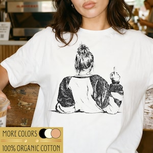 Könnte beinhalten: Weißes T-Shirt mit einer Schwarz-Weiß-Illustration einer Person von hinten, die den Mittelfinger zeigt. Das Shirt hat ein Raglanärmel-Design. Der Text "MORE COLORS" und "100% ORGANIC COTTON" sind unten aufgedruckt.