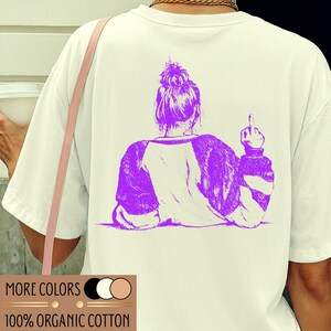 Könnte beinhalten: Cremefarbenes T-Shirt mit einem lila Aufdruck auf dem Rücken, der eine Person mit erhobenem Mittelfinger zeigt. Das Shirt hat einen Rundhalsausschnitt und lange Ärmel. Der Text "MORE COLORS" und "100% ORGANIC COTTON" ist unten aufgedruckt.