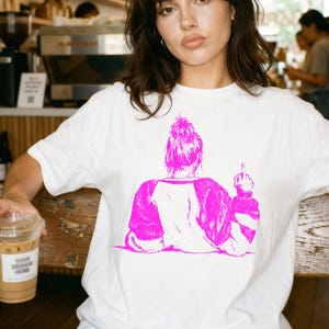 Könnte beinhalten: Weißes T-Shirt mit einer rosa Grafik einer Person von hinten, die den Mittelfinger zeigt. Das Haar der Person ist zu einem Dutt gebunden. Das Shirt hat rosa Ärmel und eine rosa Grafik auf dem Rücken.