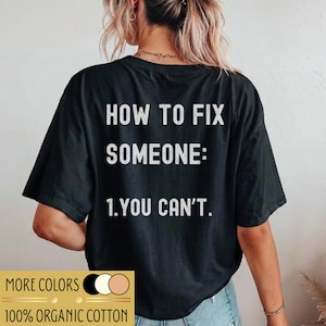 Könnte beinhalten: Ein schwarzes T-Shirt mit kurzen Ärmeln und weißem Text auf der Rückseite: HOW TO FIX SOMEONE: 1. YOU CAN'T. Das Shirt besteht aus 100 % Bio-Baumwolle und wird von hinten gezeigt, wobei die aufgedruckte Botschaft in einer serifenlosen Schrift erscheint.