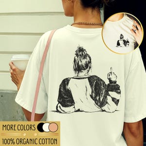 May include: Off-white t-shirt with a black and white illustration on the back of a person with their back turned, flipping the middle finger. The shirt also has the text "MORE COLORS" and "100% ORGANIC COTTON".