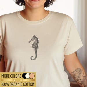 Könnte beinhalten: Cremefarbenes T-Shirt mit einer detaillierten schwarz-grauen Seepferdchen-Illustration. Das Shirt hat einen Rundhalsausschnitt und kurze Ärmel. Der Text "MORE COLORS" und "100% ORGANIC COTTON" ist unten aufgedruckt.