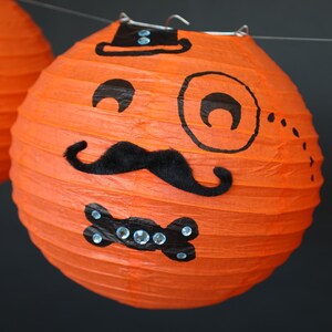 May include: Orange paper lantern with a black mustache, bow tie, and monocle drawn on it. The lantern has a black top hat drawn on it with two white dots.