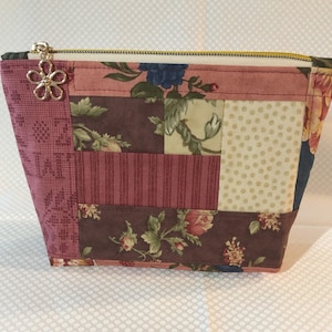 May include: A pink and brown patchwork zippered pouch with a floral design. The pouch has a gold zipper and a gold flower charm.