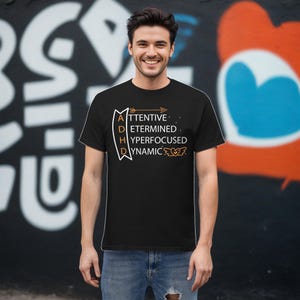 May include: A black t-shirt with a white graphic design that says "ADHD: Attentive, Determined, Hyperfocused, Dynamic". The graphic is in the shape of a ribbon with an arrow at the top.