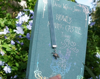 May include: A teal book titled "Howl's Moving Castle Trilogy" with silver lettering and a matching velvet bookmark. The bookmark has a silver charm. The book is held in front of a background of green foliage and blue flowers.