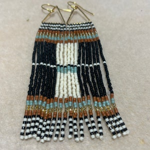 May include: A pair of long, beaded earrings with a geometric pattern in black, white, brown, and light blue. The earrings are made with small, seed beads and have a gold triangle-shaped top.