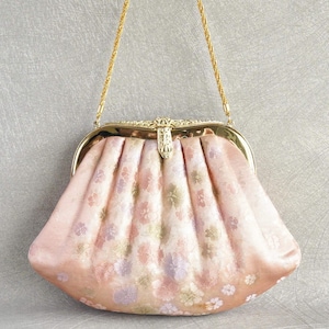 以下が含まれることがあります： ゴールドチェーンストラップが付いた、ヴィンテージスタイルの薄いピンク色のハンドバッグ。 バッグは花柄で、装飾的な留め金が付いています。 バッグのプリーツデザインは、そのクラシックな魅力を高めています。 スタイリッシュなアクセサリーです。