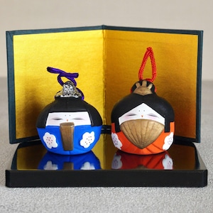 May include: Two Japanese dolls, one blue and one orange, sit on a black mirrored surface. The dolls have black hair and painted faces. They are displayed in a black box with a gold interior.