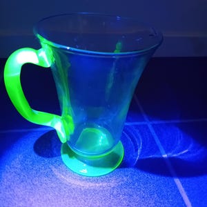 May include: A clear glass mug with a green handle and base. The mug has a flared shape and is illuminated with a blue light, creating a glowing effect on the green parts. The mug is sitting on a blue surface.