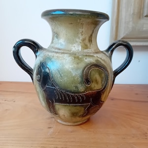 Peut inclure: Un petit vase en céramique vert et brun avec deux anses noires. Le vase présente un motif animal noir stylisé sur le devant.