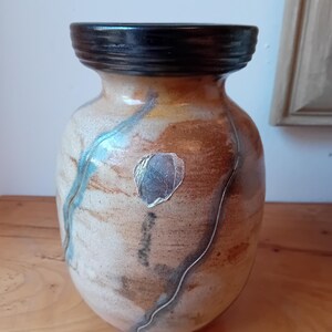 Peut inclure: Un vase en céramique avec un glaçage brun, blanc et bleu. Le vase a un bord noir et un motif de feuille d'argent.
