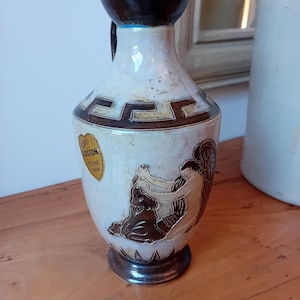 Peut inclure: Vase en céramique vintage avec un corps crème, un bord et une base marron foncé. Le vase présente un motif de clé grecque et une étiquette indiquant "Gres Losson Tourne Main". Une illustration de personnages est sur le côté.