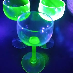 May include: Three clear glass drinking glasses with a green tint. The glasses are illuminated with a black light, causing them to glow a bright green.
