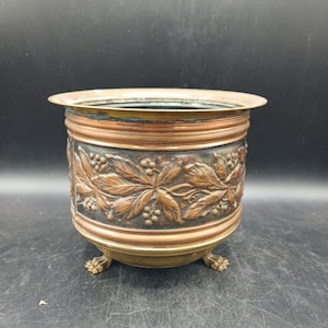 Pode incluir: Um vaso de cobre redondo com um design em relevo de folhas e bagas à volta do centro. O recipiente tem um rebordo largo e liso, uma pátina escura nos detalhes em relevo e assenta sobre três pequenos pés metálicos em forma de garra.