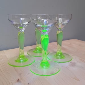 May include: Four clear glass cocktail glasses with neon green stems and bases. The stems are a vibrant green, and the bases are circular. The glasses are arranged on a light-colored wooden surface.