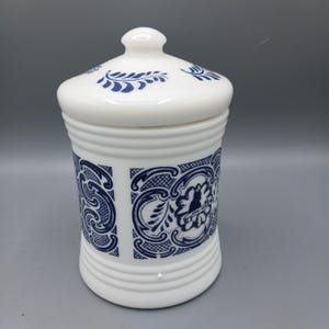 Può includere: Un barattolo in vetro opalino bianco con coperchio abbinato, caratterizzato da un motivo decorativo blu scuro con volute ornamentali e una silhouette centrale con la scritta "ANNO 1804". Il contenitore cilindrico presenta dettagli a coste.