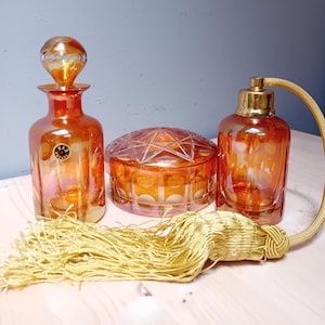 May include: An orange glass vanity set with a perfume bottle, a round lidded jar, and a perfume atomizer. The set includes a gold tassel. The glass has a reflective, iridescent finish.