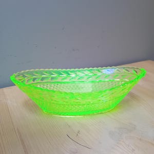 May include: A neon green, oval-shaped glass bowl. The bowl has a textured surface with a diamond pattern and a decorative leaf design around the rim. The bowl is sitting on a light-colored wooden surface.