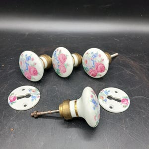 以下が含まれることがあります： 真鍮金具付きのヴィンテージ白磁ドアノブのセット。各ノブは、ピンクと青のバラのフローラルデザインが特徴です。キーホールカバーも含まれています。