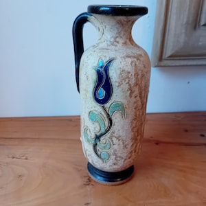 Peut inclure: Un vase en céramique blanche avec une anse noire et un motif floral bleu et vert. Le vase a une surface texturée et une base noire.