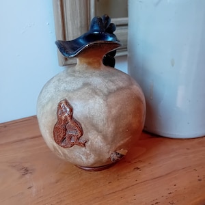 Peut inclure: Un pichet en céramique avec un glaçage brun et un bord noir. Le pichet a un corps rond et un petit bec verseur. Un motif brun d'un oiseau est sur le côté du pichet.