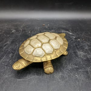 Pode incluir: Uma estatueta de tartaruga em latão com um corpo de metal dourado polido e um casco decorado com painéis incrustados de madrepérola branca. A tartaruga tem uma cabeça detalhada, quatro patas e um padrão de casco texturizado com segmentos geométricos.