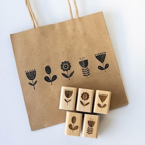 Pode incluir: Um conjunto de seis carimbos de madeira com diferentes desenhos florais. Os carimbos estão dispostos em uma fila sobre um saco de papel castanho com alça. Os carimbos são preto e branco e têm um estilo simples e minimalista.