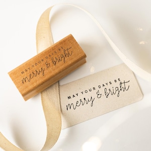 Op de afbeelding: Een houten stempel met de tekst "MAY YOUR DAYS BE merry & bright" ligt naast een bijpassend label. Een crèmekleurig lint is om de stempel en het label gekruld. De achtergrond is wit met zachte, vage highlights.