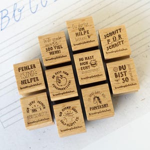 Könnte beinhalten: Eine Ansammlung kleiner, quadratischer Holzstempel mit verschiedenen deutschen Sätzen und Illustrationen. Die Stempel sind auf einem Blatt Papier mit handgeschriebenen Kursivbuchstaben und Notenlinien angeordnet. Zu den Sätzen gehören "Fehler sind Helfer" und "Du bist so".