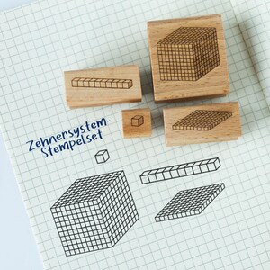 Op de afbeelding: Een set houten stempels met geometrische ontwerpen, waaronder kubussen, rechthoekige prisma's en vierkanten. De stempels zijn gerangschikt op geruit papier, met de blauwe tekst "Zehnersystem-Stempelset".
