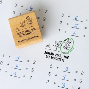 Op de afbeelding: Een houten stempel met de tekst "SCHAU MAL, WIE DU W&Auml;CHST! kreativp&uuml;nktchen" en een bloemenontwerp, geplaatst op een werkblad met handgeschreven wiskundeproblemen. Het werkblad bevat optelproblemen.