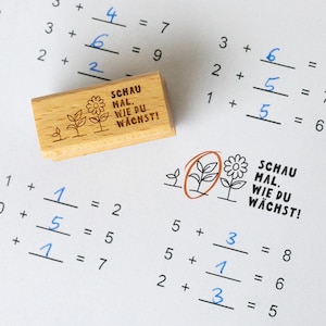 Op de afbeelding: Een houten stempel met de tekst "SCHAU MAL, WIE DU WÄCHST!" en plantillustraties ligt naast een wiskundewoordenblad met optelsommen. De stempel is rechthoekig en lichtbruin.