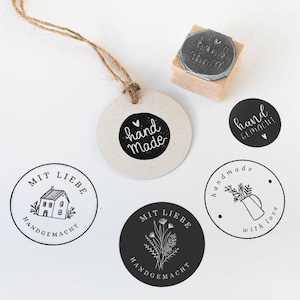 May include: A selection of handmade craft supplies, including round tags, stickers, and a wooden stamp. The tags and stickers feature black and white designs with phrases like "hand made" and floral illustrations. A natural twine is attached to one tag.