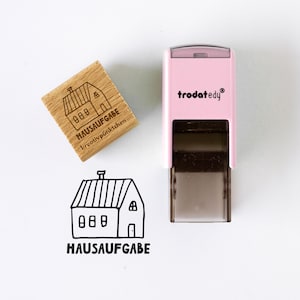 Può includere: Un timbro autoinchiostrante rosa chiaro con l'etichetta trodatedy e un timbro quadrato in legno con l'illustrazione di una casa e il testo HAUSAUFGABE e kreativpünktchen. Sotto, un'impronta a inchiostro nero mostra la casa e HAUSAUFGABE.
