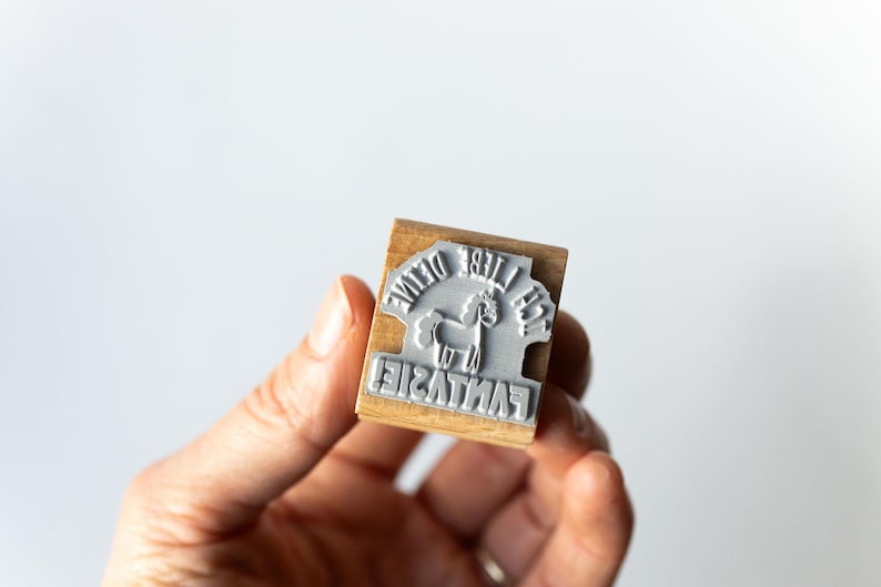 May include: A wooden stamp with a gray rubber stamp face. The stamp reads "The Divine Fantasie" around a graphic of a unicorn. The stamp is held in a person's hand against a white background.