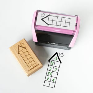 Può includere: Un timbro rosa e nero con un inserto bianco con un design a freccia e griglia neri. È presente anche un timbro in legno con un design simile e la scritta "kreativpünktchen". Una freccia disegnata a mano con numeri è accanto ai timbri.