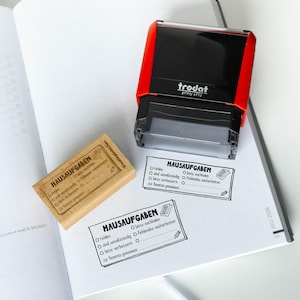 Könnte beinhalten: Ein rot-schwarzer Trodat Printy 4913 Selbstfärbestempel, ein Holzstempel und zwei schwarze Stempelabdrücke auf Papier. Die Stempel zeigen den Text HAUSAUFGABEN, eine Checkliste mit Kästchen sowie Symbole eines Buches und eines Bleistifts.