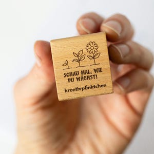 Op de afbeelding: Een kleine, vierkante houten stempel met een bloemenontwerp en Duitse tekst. De stempel toont de groei van een plant, van een scheut tot een bloem. De tekst luidt "SCHAU MAL, WIE DU W&Auml;CHST!" en "kreativp&uuml;nktchen".