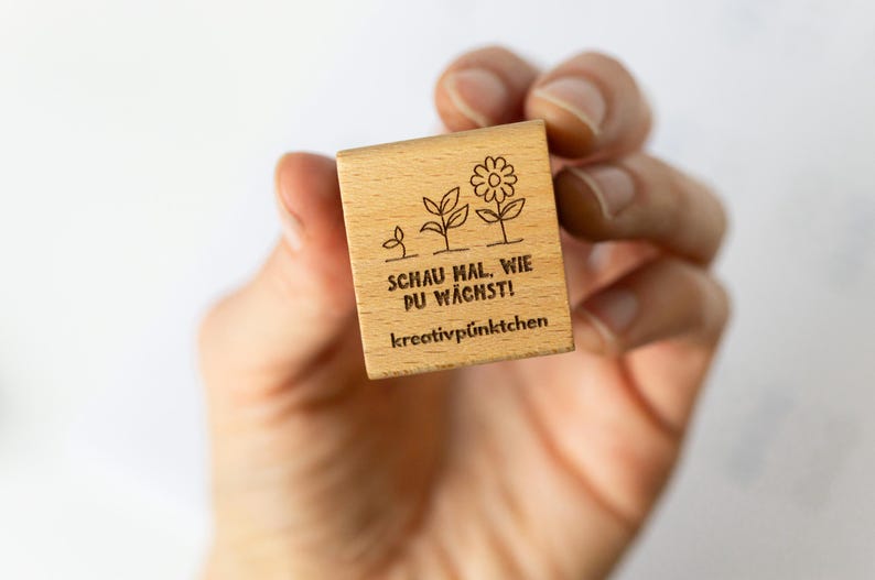 Op de afbeelding: Een kleine, vierkante houten stempel met een bloemenontwerp en Duitse tekst. De stempel toont de groei van een plant, van een scheut tot een bloem. De tekst luidt "SCHAU MAL, WIE DU W&Auml;CHST!" en "kreativp&uuml;nktchen".