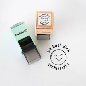Op de afbeelding: Twee stempels, één mintgroen en één beige, met de tekst "Du hast dich verbessert!" en een lachend gezicht. De mintgroene stempel heeft de merknaam "trodat edy" erop.