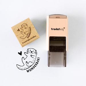 May include: A wooden stamp with a cute otter design, a heart, and the words "WUNDERBAR!" and "kreativpünktchen". A self-inking stamp with the brand name "trodat edy" is also present. The otter illustration is in black.