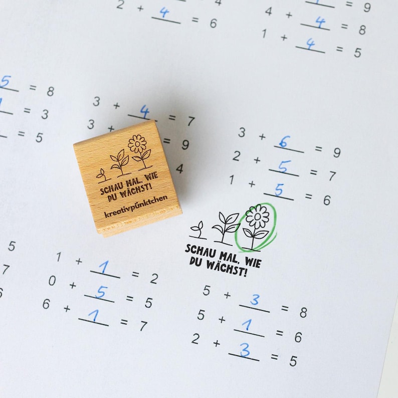 Op de afbeelding: Een houten stempel met de tekst "SCHAU MAL, WIE DU W&Auml;CHST! kreativp&uuml;nktchen" en een bloemenontwerp, geplaatst op een werkblad met handgeschreven wiskundeproblemen. Het werkblad bevat optelproblemen.